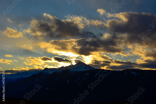 The sun sets behind the clouds in the shadow above the snowy evening mountains