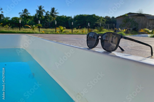 Black sunglasses on the edge of the blue swimming pool in secluded spot with the fence and greenery in the background blurred in the radiant light of the summer sun.