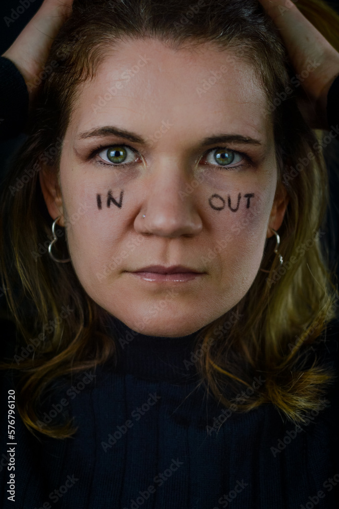 Fototapeta premium making decisions - portrait of a beautiful blonde woman with the words in and out written on her cheeks and looking intently in uncertainty