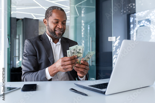 Successful boss banker inside office sitting at workplace with laptop, happy african american investor holding cash money dollars in hands, man counting profit income happy with achievement result.