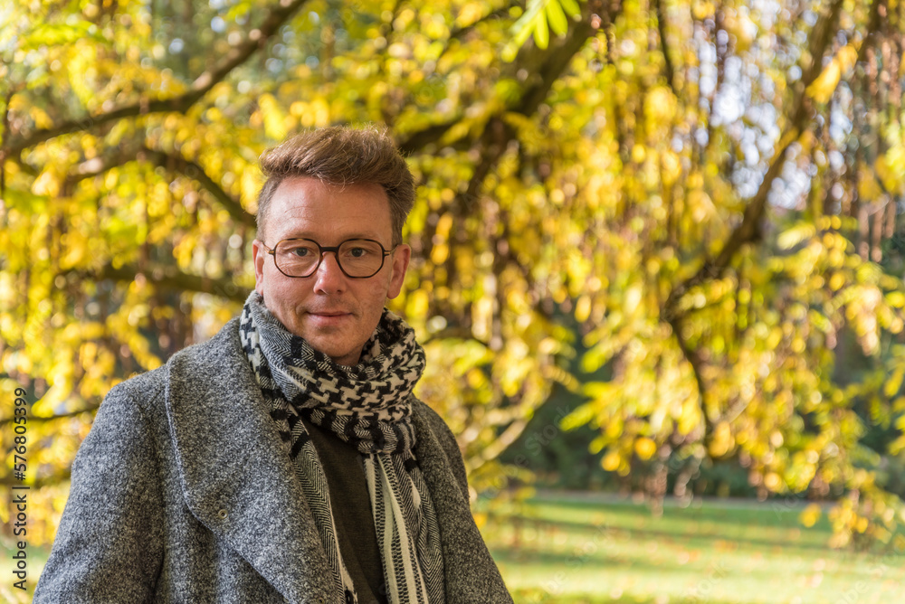 Caucasian male posing in front of a tree with leaves in autumn colors and branches in a public park in Berlin, Germany Caucasian male posing in front of a tree with leaves in autumn colors and branches in a public park in Berlin, Germany