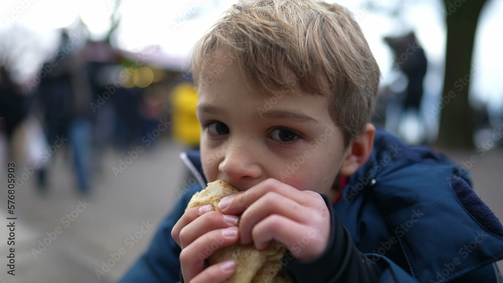 Small boy eating pancake outside in city street during winter season. Hungry child eating food during december festivities Small boy eating pancake outside in city street during winter season. Hungry child eating food during december festivities