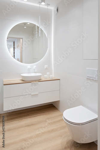 Luxury bathroom with round mirror with led lights, stylish washbasin and wooden floor. Modern interior of bathroom with wc and bath. Vertical.