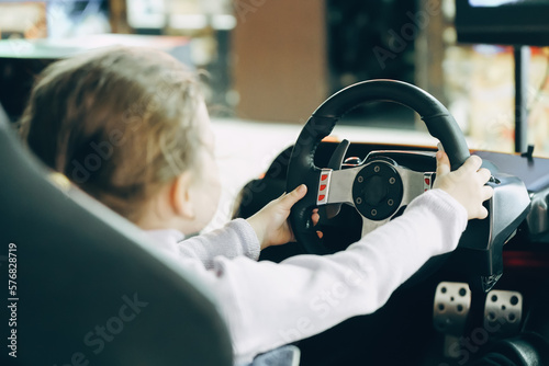 A little girl in an entertainment center with slot machines sits in a car game simulator and holds the steering wheel with her hands