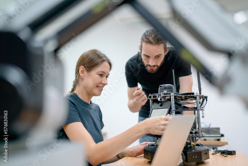 Two Caucasian professional technician or engineer workers sit in workplace and help to check and maintenance small robotic machine on table and use laptop to support.