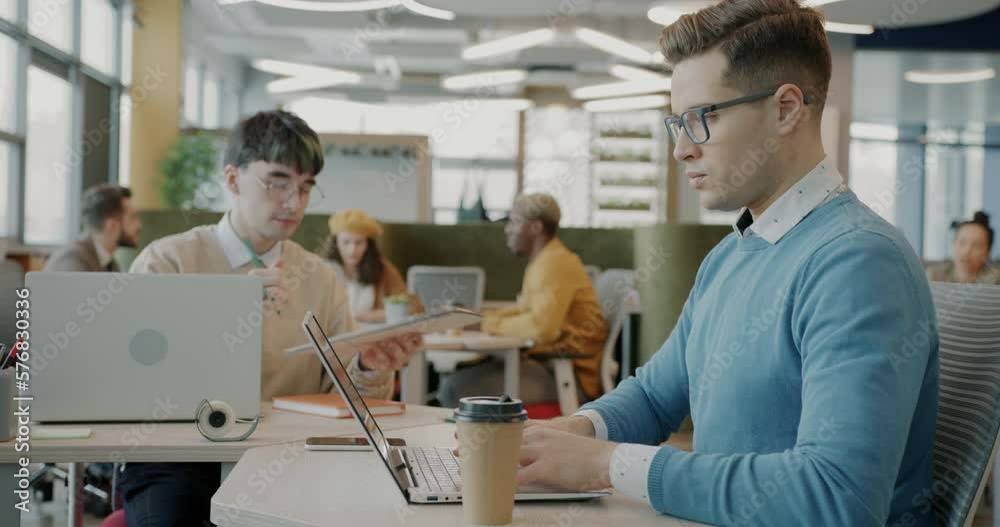 Ambitious businessman working with laptop computer and talking to male colleague in creative coworking space. Business and job concept. Ambitious businessman working with laptop computer and talking to male colleague in creative coworking space. Business and job concept.