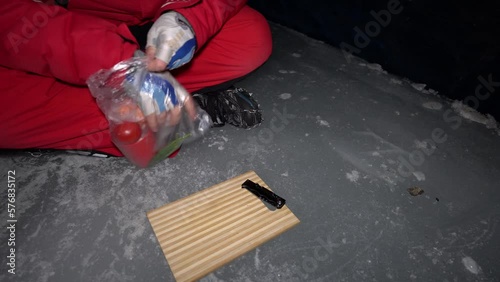 A guy cooks food on gas inside an ice cave. The climber lights the burner, cuts sausage and tomato. Breaks the eggs and fry in a frying pan. Steam from food. The ice wall is blue and the floor is gray photo