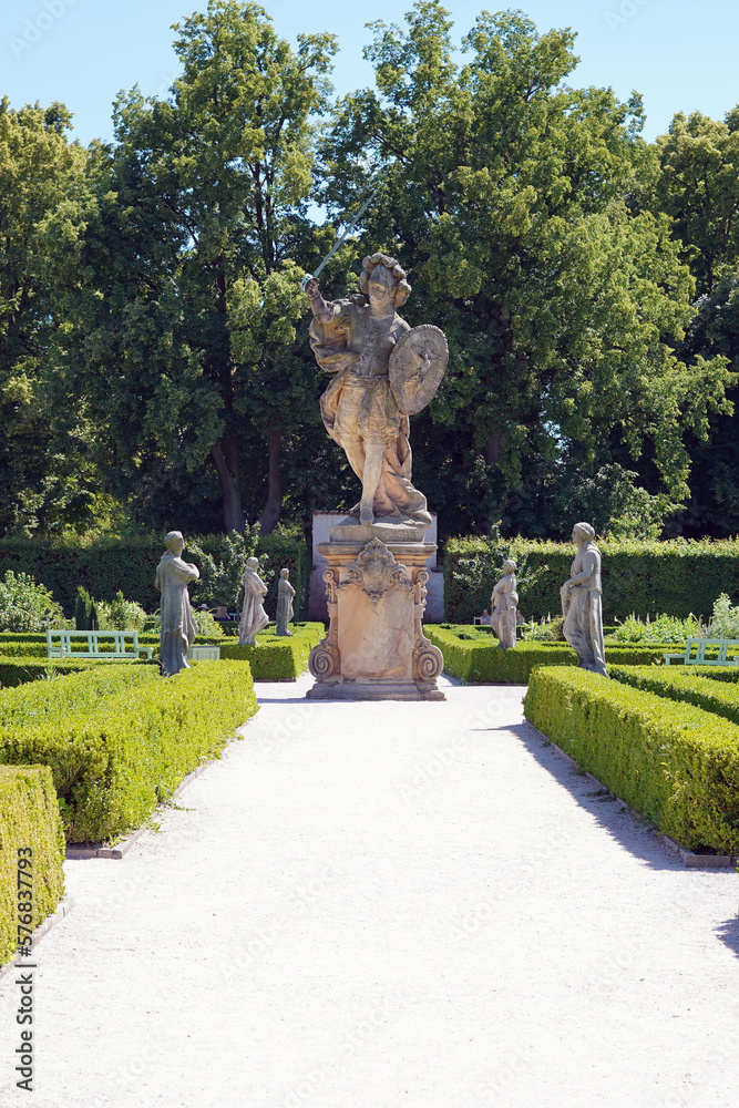 Statue in the castle garden in superhuman size. A sandy path lined with bushes leads to it.