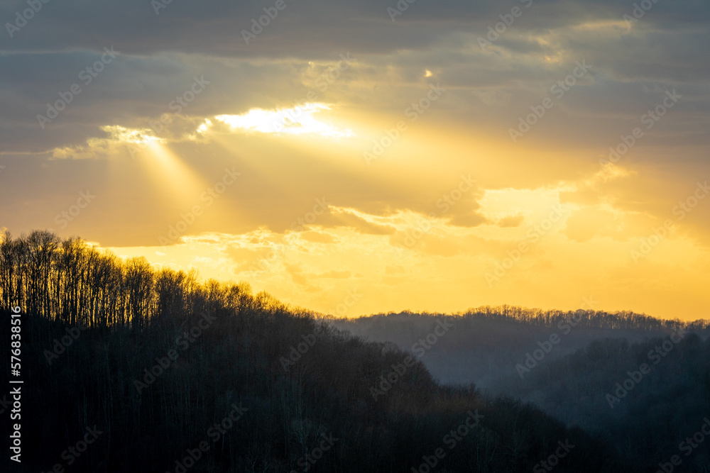 Yellow sun rays shining through dark clouds over forest silhouette