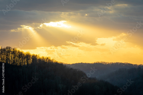 Yellow sun rays shining through dark clouds over forest silhouette