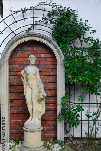 posąg kobiety w ogrodzie na tle ceglanej ściany, posąg kobiety z piaskowca otoczony pnączami, statue of a woman in garden, garden statue of a woman surrounded by green climber, brick wall in garden