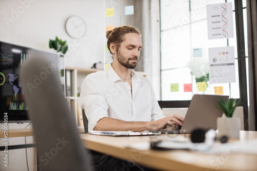 Portrait of attractive young business man sitting at office desk and working with laptop. Caucasian guy with beard having video conference. Business meeting online.