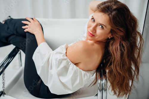 a beautiful woman with long hair in a white top and black pants lies on a chair.