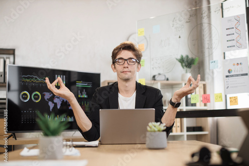 Relaxed confident caucasian man in suit sitting on workplace with modern laptop and meditating with closed eyes. Office worker relief stress at work with accomplished fingers.