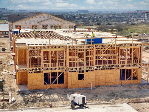 New Apartment Building Complex Under Construction - Workers Constructing The Floors And Roof With Wood Frame And Plywood On A Sunny Day With Blue Skies And Equipment On The Street