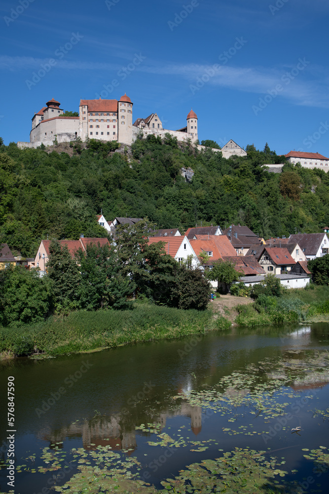 Landscape shot of Harburg Castle in Bavaria, which stands on the bank of a river. The castle stands on a hill above a village. In between is a forest. The blue sky above.