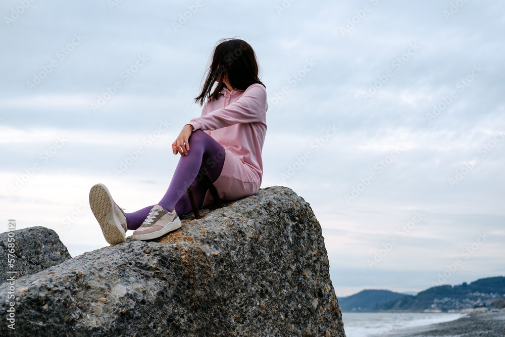 A girl on the beach is sitting on a big rock and looking at the sea