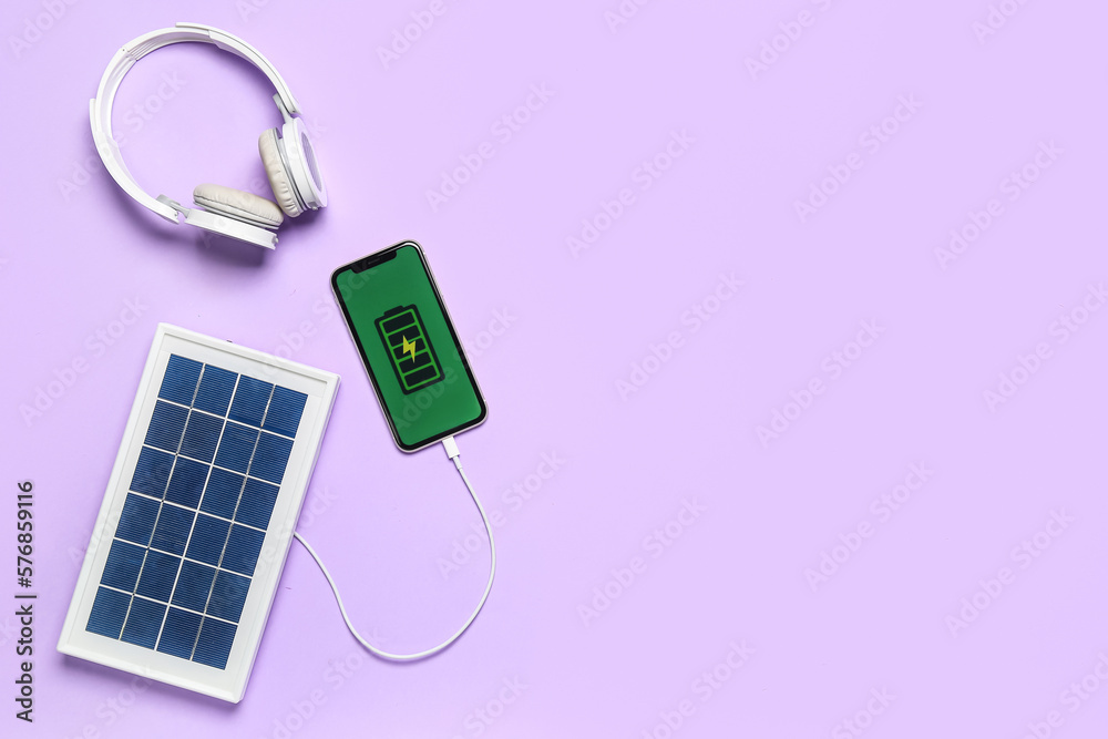 Portable solar panel charging mobile phone and headphones on lilac background