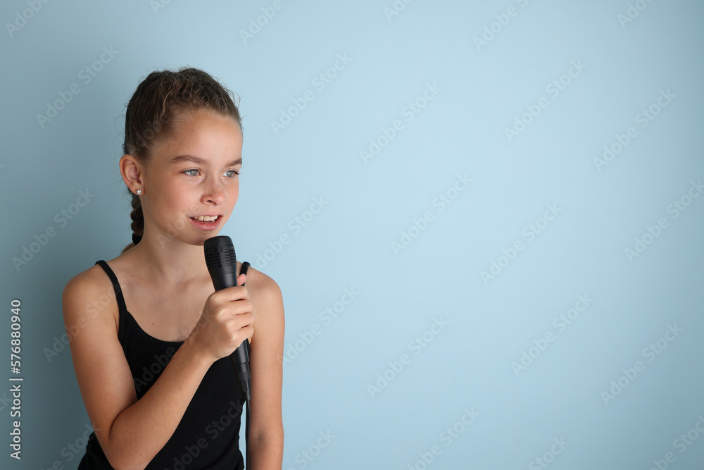 Portrait of a cute teenage girl singing, using microphone. Teenage girl in black t-shirt on an isolated blue background. Karaoke for kids, home entertainment for kids. Singer singing with microphone. Portrait of a cute teenage girl singing, using microphone. Teenage girl in black t-shirt on an isolated blue background. Karaoke for kids, home entertainment for kids. Singer singing with microphone.