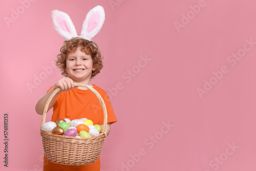 Portrait of happy boy in cute bunny ears headband holding wicker basket with Easter eggs on pink background. Space for text