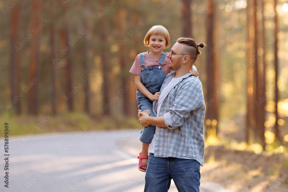 Fototapeta premium Dad holds a little daughter in his arms, they laugh merrily walking along a forest road among tall pines. Family walk in the forest at sunset, man and little girl.