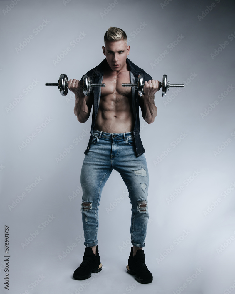 Powerful full-length portrait of muscular sexy man in jeans with dumbbells at white background. Handsome fitness male model with gym equipment in studio. Powerful full-length portrait of muscular sexy man in jeans with dumbbells at white background. Handsome fitness male model with gym equipment in studio.