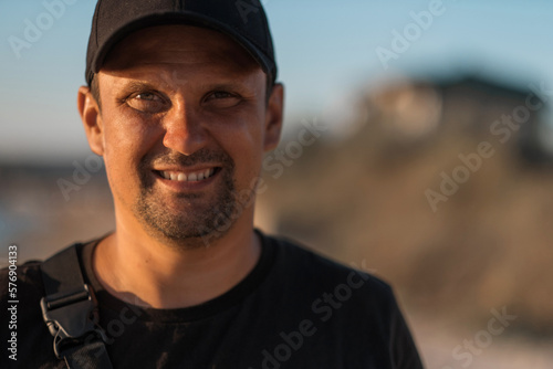 A handsome brunette man in a black t-shirt smiles and looks at the camera against the backdrop of mountains and a blue sky. High quality photo. © aaalll3110