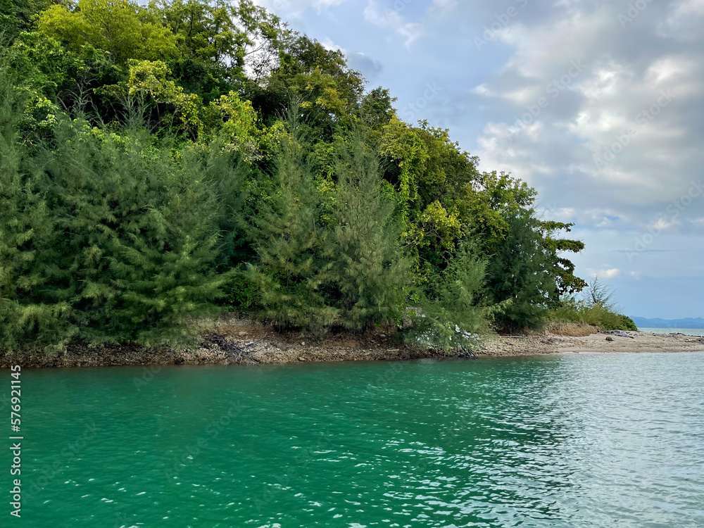 Tropical island. Bright emerald color calm water in the foreground. Intense juicy green foliage of dense forest covering a hill. Sandy shore. Blue sky with clouds. Perfect landscape. View from the sea