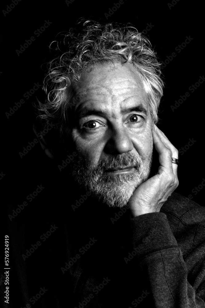 Black and white portrait of an attractive older Caucasian man with grey hair and beard. Looking towards the camera, leaning on his hand, relaxed and thoughtful expression.