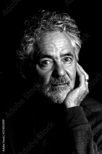 Black and white portrait of an attractive older Caucasian man with grey hair and beard. Looking towards the camera, leaning on his hand, relaxed and thoughtful expression.