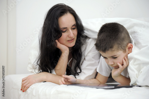 mom and son 7 years old lying under blanket on white sheets boy looking at tablet playing game mom turned to face him love for children care online learning english course learning technology getting