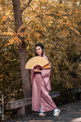 Japanese geisha in a traditional kimano with a fan and armed with a katana sword in a beautiful garden. A girl from medieval Asia. Reconstruction of cultural heritage. Culture in Japan.