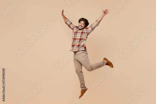 Full body side view young Indian man wear brown shirt casual clothes jump high with outstretched hands arms isolated on plain pastel light beige background studio portrait. People lifestyle concept.