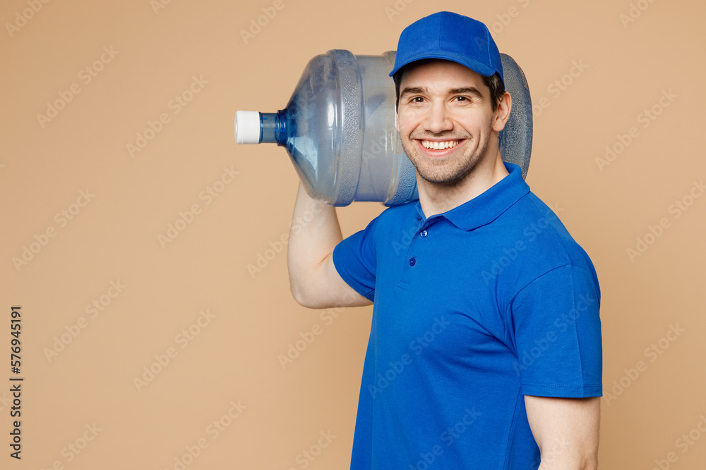 Side view fun delivery guy employee man wearing blue cap t-shirt uniform workwear working as dealer courier hold big water bottle on shoulder isolated on plain light beige background. Service concept. Side view fun delivery guy employee man wearing blue cap t-shirt uniform workwear working as dealer courier hold big water bottle on shoulder isolated on plain light beige background. Service concept.