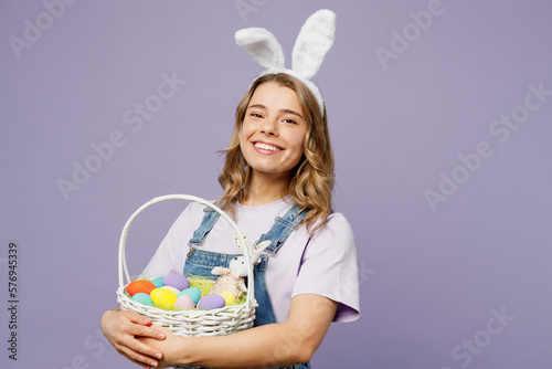Leinwand Poster Young smiling cheerful funny woman wearing casual clothes bunny rabbit ears holding wicker basket colorful eggs looking camera isolated on plain pastel purple background studio