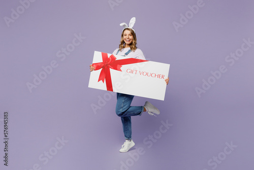 Full body young woman wear casual clothes bunny rabbit ears hold store big gift certificate coupon voucher card isolated on plain pastel light purple background studio. Lifestyle Happy Easter concept.