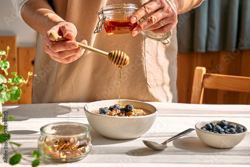 Woman preparing healthy dieting vegan nutritious breakfast. Female hand pouring honey in the bowl with oatmeal porridge with walnuts and blueberries.