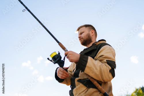 Close-up low-angle view of bearded fisherman holding casting rod wearing raincoat standing on bank waiting for bites on water river at summer day. Concept of lifestyle, leisure activity on nature.