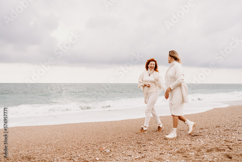 Women sea walk friendship spring. Two girlfriends, redhead and blonde, middle-aged walk along the sandy beach of the sea, dressed in white clothes. Against the backdrop of a cloudy sky and the winter