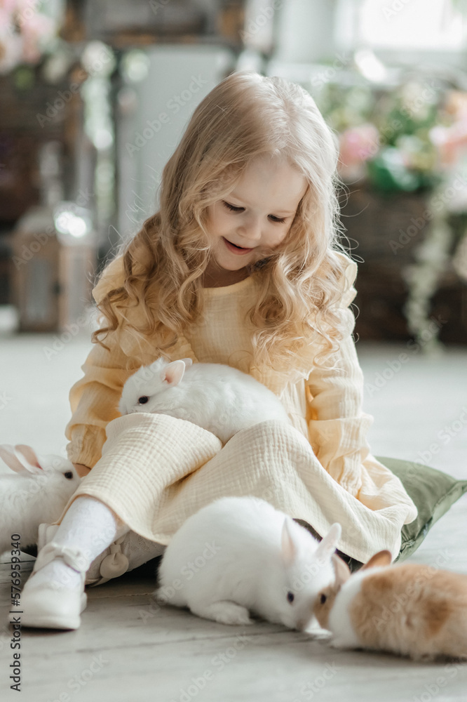 A little smiling girl in a yellow dress is playing on the floor in a room with rabbits. A baby and a rabbit. The living room is decorated for Easter. A little smiling girl in a yellow dress is playing on the floor in a room with rabbits. A baby and a rabbit. The living room is decorated for Easter.