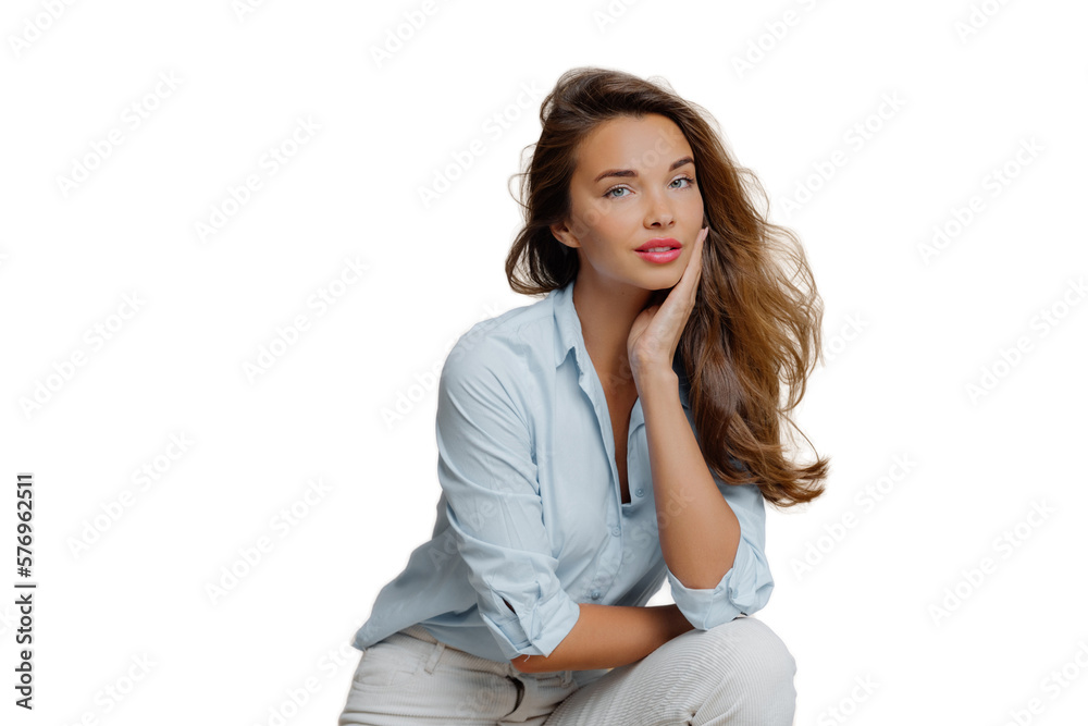 Portrait of attractive young woman sits against brown background, touches cheek, dressed in fashionbale outfit, has thoughtful look at camera, demonstrates her beauty. People and style concept Portrait of attractive young woman sits against brown background, touches cheek, dressed in fashionbale outfit, has thoughtful look at camera, demonstrates her beauty. People and style concept