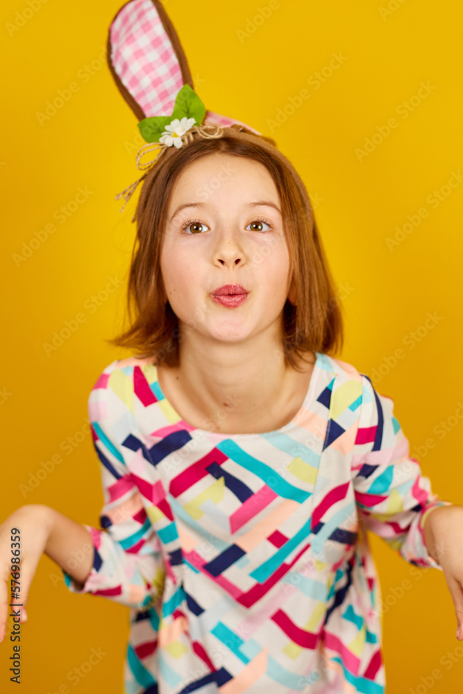 Happy playful teenager girl wearing bunny ears on a bright yellow studio background, celebrating Easter in style.