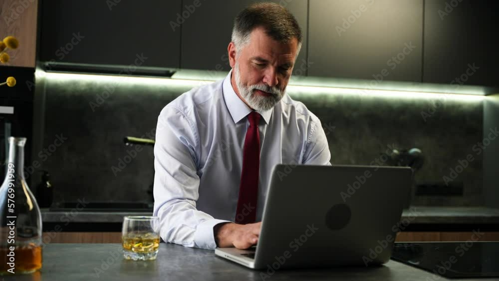 Mature man working on laptop and drinking whiskey at home kitchen. Businessman having alcohol while using notebook Mature man working on laptop and drinking whiskey at home kitchen. Businessman having alcohol while using notebook