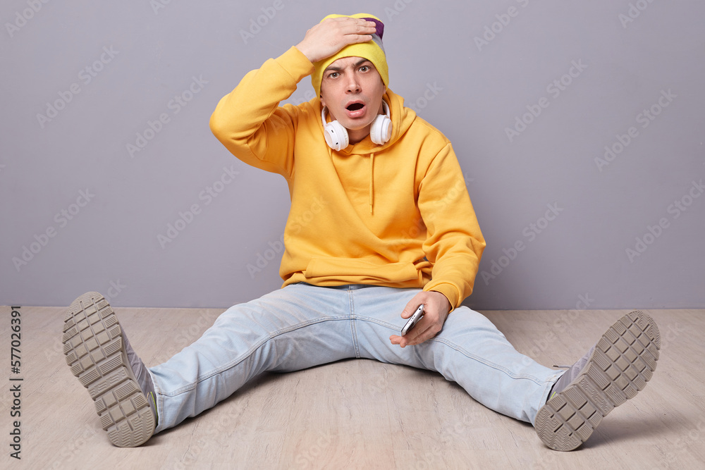 Portrait of shocked disappointed man wearing beanie hat, yellow hoodie and jeans posing against gray wall, sitting on floor, making facepalm gesture, forgot something important. Portrait of shocked disappointed man wearing beanie hat, yellow hoodie and jeans posing against gray wall, sitting on floor, making facepalm gesture, forgot something important.