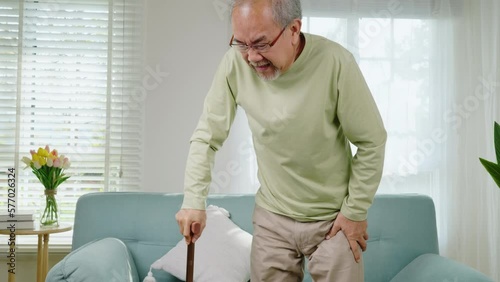Asian senior old man with eyeglasses type to standing up from sofa with walking cane stick to walk at home, Elderly suffering from knee pain ache holding handle of cane, retirement medical healthcare