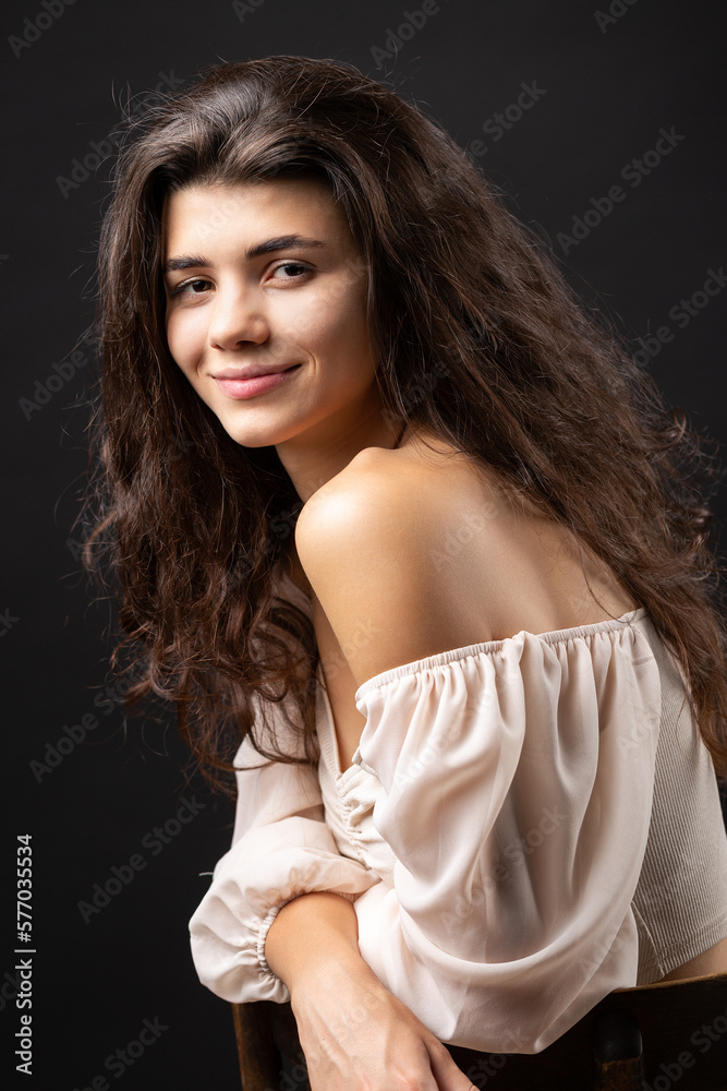 Classic studio portrait of a young brunette in a white loose top, who is sitting on a chair against a black background.