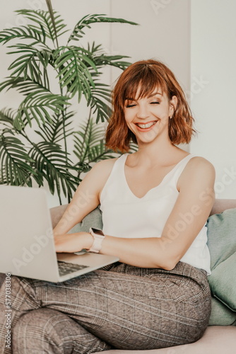 Brunette macbook sofa. Attractive young woman working on a laptop while sitting on the couch at home. She is wearing a light blouse and trousers.