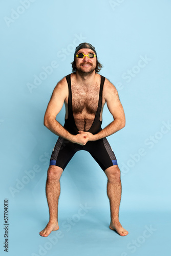 Comic portrait of funny bearded man in swimsuit and swimming cap and goggles having fun isolated over blue background. Concept of sport, active lifestyle and humor