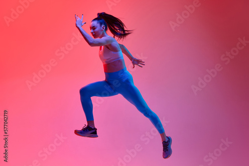 Full-length image of young girl, professional athlete, runner in motion, training over pink studio background in neon light. Concept of sportive lifestyle, health, endurance, action and motion. Ad