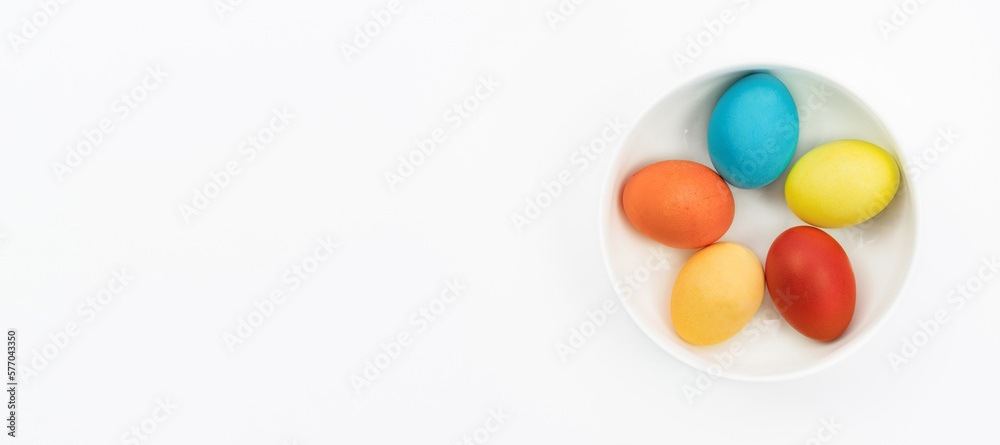 Easter eggs lie in a bowl on a white background with copy space. Easter banner in a minimalistic style. Traditional Easter eggs painted by kids in a white plate top view.